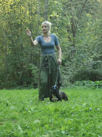 Au milieu d'une prairie, l'éducatrice explique d'un geste de la main, un bouledogue français muselé posté au calme à ses pieds.