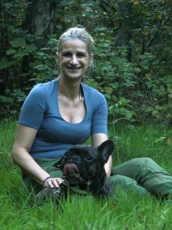 L'éducatrice, assise dans l'herbe d'un sous-bois, sourit à l'objectif, un bouledogue français bringé calé contre elle.