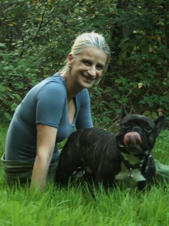 Assise dans l'herbe d'un sous-bois, l'éducatrice sourit à côté d'un bouledogue français bringé qui se lèche la truffe.